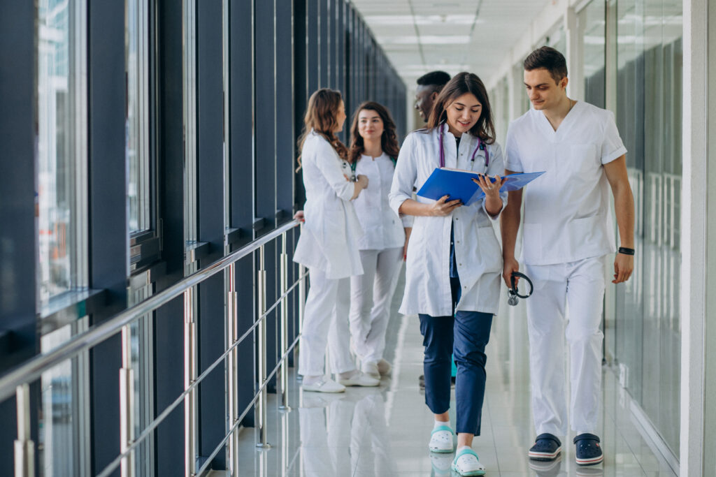 Symbolbild auf dem mehrere Ärzt*innen in einem Krankenhausflur zu sehen sind. Zwei Ärzt*innen laufen in Richtung Kamera und schauen sich gemeinsam eine Krankenakte an.